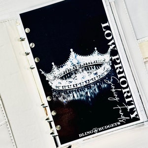 May include: A white planner with a clear plastic cover. The cover has a black background with a silver crown and the text "LOW PRIORITY Sipping Funds" and "BLING & BUDGETS".