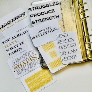 May include: A collection of white cards with motivational phrases in black and gold lettering. Phrases include "Manifest that shit," "Struggles Produce Strength," and "Mindset is Everything." The cards are arranged near a gold planner.