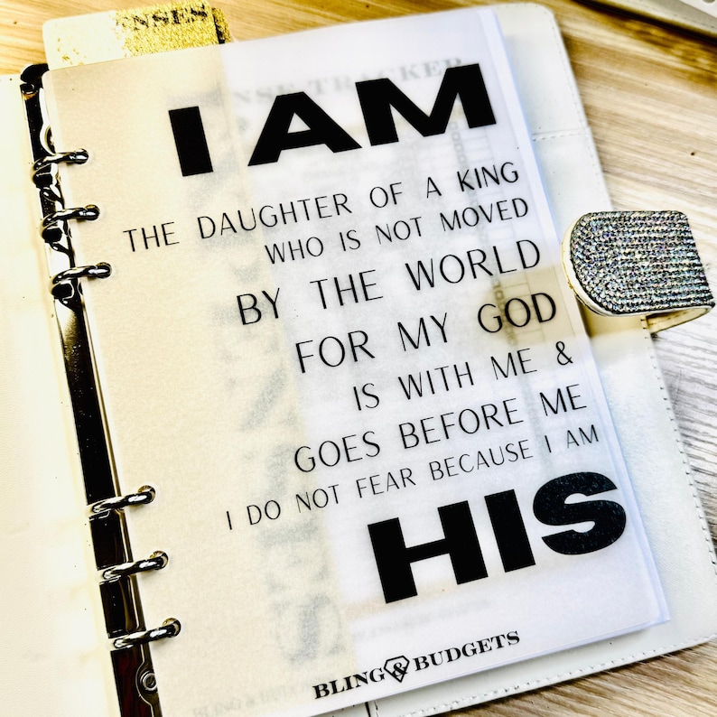 Op de afbeelding: Een witte planner met een transparante overlay met de tekst "I AM THE DAUGHTER OF A KING WHO IS NOT MOVED BY THE WORLD FOR MY GOD IS WITH ME & GOES BEFORE ME I DO NOT FEAR BECAUSE I AM HIS". De planner heeft een zilveren ringband en een strass sluiting.