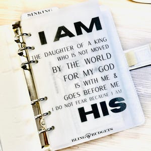 Op de afbeelding: Een witte planner met een transparante overlay met de tekst "I AM THE DAUGHTER OF A KING". De planner heeft zilveren metalen ringen en een witte band. De woorden "BLING BUDGETS" staan onderaan.