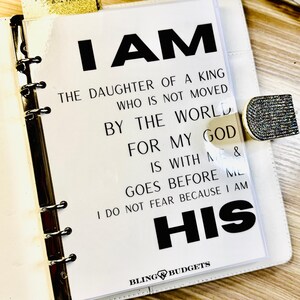 Op de afbeelding: Witte planner met de tekst "I AM THE DAUGHTER OF A KING WHO IS NOT MOVED BY THE WORLD FOR MY GOD IS WITH ME & GOES BEFORE ME I DO NOT FEAR BECAUSE I AM HIS" in het zwart. De planner heeft een zilveren binder en een sprankelende sluiting.