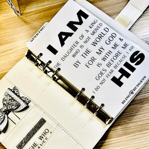 Op de afbeelding: Witte kunstleren planner met zilveren accenten en de tekst "I AM THE DAUGHTER OF A KING" gedrukt op een pagina. De planner heeft een strik en een vakje met de woorden "SHE WHO AVEC". De planner is open.