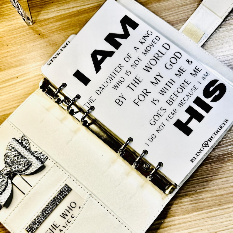 Op de afbeelding: Witte kunstleren planner met zilveren accenten en de tekst "I AM THE DAUGHTER OF A KING" gedrukt op een pagina. De planner heeft een strik en een vakje met de woorden "SHE WHO AVEC". De planner is open.