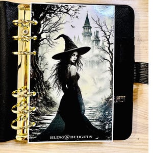 May include: Black planner with gold-colored rings and a decorative insert featuring a witch in a black dress and hat, with a castle in the background. The text "BLING & BUDGETS" is at the bottom.