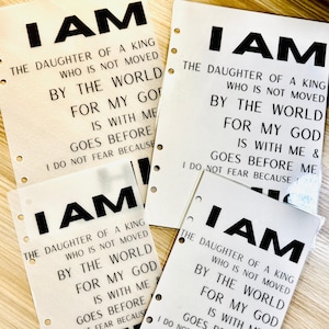 Op de afbeelding: Verschillende witte papieren vellen met de woorden "I AM" in vette zwarte letters bovenaan. De tekst hieronder luidt: "THE DAUGHTER OF A KING WHO IS NOT MOVED BY THE WORLD FOR MY GOD IS WITH ME & GOES BEFORE ME I DO NOT FEAR BECAUSE."