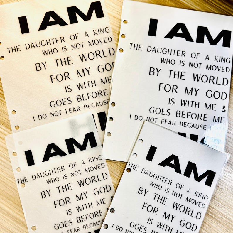 Op de afbeelding: Verschillende witte papieren vellen met de woorden "I AM" in vette zwarte letters bovenaan. De tekst hieronder luidt: "THE DAUGHTER OF A KING WHO IS NOT MOVED BY THE WORLD FOR MY GOD IS WITH ME & GOES BEFORE ME I DO NOT FEAR BECAUSE."
