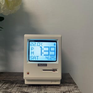 Puede incluir: Un reloj digital blanco con forma de monitor de ordenador antiguo. La pantalla azul muestra la hora, la fecha y la temperatura. El reloj tiene la palabra "Maclock" y el logotipo de Apple.
