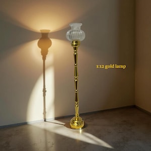 May include: A gold floor lamp with a clear, ribbed glass shade. The lamp has a slender, ornate stem and a round base. The text "1:12 gold lamp" is visible on the right side of the image. The lamp casts a shadow on the wall.