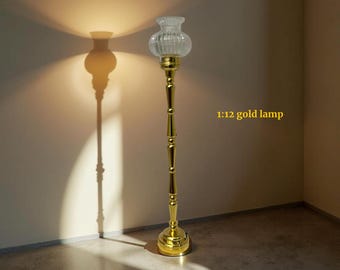 May include: A gold-colored floor lamp with a clear, ribbed glass shade. The lamp has a slender, ornate stem and a round base. The text "1:12 gold lamp" is visible on the right side of the image. The lamp casts a shadow on the wall.