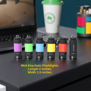 May include: Six mini keychain flashlights in a row, each with a different colour: purple, green, blue, yellow, red, and black. Each flashlight is 5.08 cm long and 3.81 cm wide, with a keyring. The text "mini-torch" is printed on each.