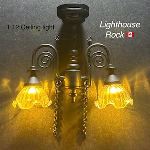 May include: A miniature black ceiling light with two lamps. The lamps have yellow glass shades and are lit. The text "1:12 Ceiling light" and "Lighthouse Rock" are visible.