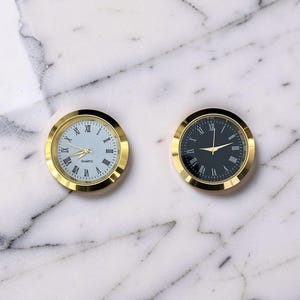May include: Two round clock inserts with gold-tone metal bezels. One clock features a white face with black Roman numerals and the word QUARTZ, while the other has a black face with white Roman numerals. Both have gold-tone hour and minute hands.