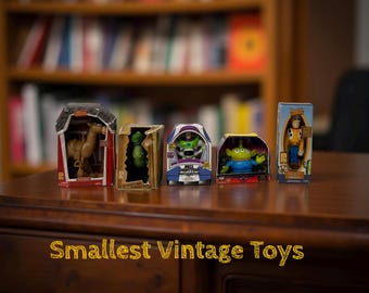 May include: A collection of five vintage toy figures in their original boxes, including a horse, dinosaur, Buzz Lightyear, an alien, and Woody. The boxes are displayed on a dark wooden surface with the text "Smallest Vintage Toys" in gold.