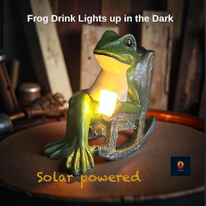 May include: A green frog figurine sitting in a rocking chair, holding a small cup that emits a warm yellow light. The text "Frog Drink Lights up in the Dark" and "Solar powered" is visible. The frog has a light-coloured chest and dark eyes.