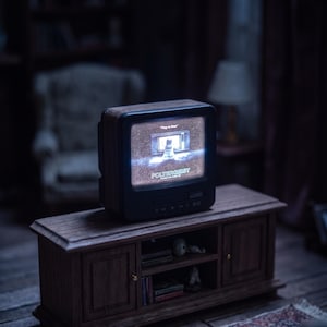 Puede incluir: Un televisor en miniatura de color negro se encuentra sobre un mueble multimedia de madera. La pantalla muestra una escena con una figura frente a un televisor y el texto "POLTERGEIST". El mueble tiene dos armarios laterales y un estante.