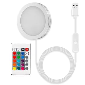 Peut inclure: Une lampe à disque LED blanche avec une bordure argentée, connectée à un câble USB et une télécommande. La télécommande propose des options de couleurs et des commandes marche/arrêt. La lumière est circulaire et émet une lueur vive.