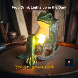 Puede incluir: Una figura de rana verde sentada en una mecedora, sosteniendo un cuadrado brillante. El texto "Frog Drink Lights up in the Dark" y "Solar powered" son visibles. Es un artículo decorativo con energía solar.