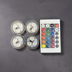 May include: Four circular LED light pucks with clear housings and three internal light diodes each, accompanied by a rectangular white remote control featuring twenty-four buttons for color selection, brightness adjustment, and lighting modes.