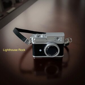 May include: A miniature black and silver camera with a black strap. The camera has a silver top and a black body with a large lens. The text "Lighthouse Rock" is visible on the left side.