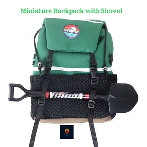 Puede incluir: Una mochila verde en miniatura con un bolsillo de malla negro y una pequeña pala adjunta. La mochila tiene rayas blancas y un parche circular con un diseño de montaña y sol. El texto "Miniature Backpack with Shovel" está en la parte superior.