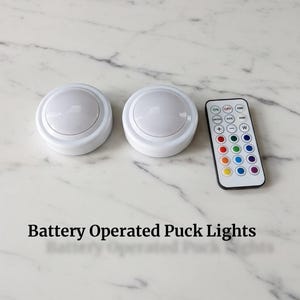May include: Two white, battery-operated puck lights with a remote control. The remote has multiple color options and buttons for on/off and dimming. The text "Battery Operated Puck Lights" is displayed below the lights.