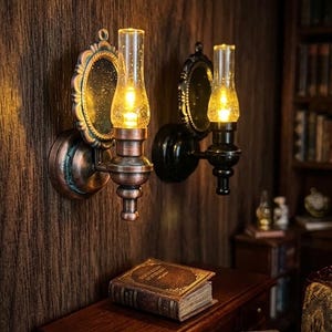 May include: Two wall-mounted sconces with copper and black finishes, each featuring a circular backplate and a clear glass chimney housing a glowing light. The fixtures are mounted on a dark wood-panelled wall above a wooden desk with a book.