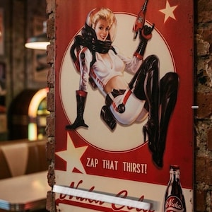 May include: A rectangular metal sign featuring a retro-style illustration of a person in a white space suit and black boots holding a ray gun. The sign includes the text ZAP THAT THIRST!, Nuka Cola, and Since 2044, with a graphic of a soda bottle.