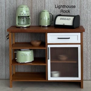 May include: A miniature kitchen set featuring a wooden cabinet with shelves and appliances. The set includes a green coffee maker, kettle, toaster, and a black bread bin with "BREAD" text. The cabinet has a white drawer and a glass-panelled door.