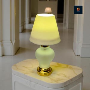 Puede incluir: Una pequeña y elegante lámpara de mesa con base de vidrio esmerilado y base de metal dorado. La pantalla es de color amarillo suave y la lámpara está sobre una mesa con tapa de mármol. La lámpara está encendida, emitiendo un brillo cálido.