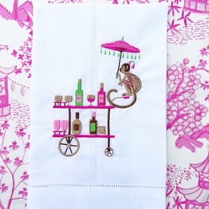 May include: White embroidered towel featuring a monkey with a pink umbrella, pushing a brown bar cart with bottles and glasses. The cart has brown wheels and pink shelves. The background is a pink and white floral pattern.