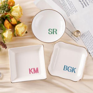 May include: Three white ceramic dishes with gold trim. The dishes are square and round. Each dish has a different set of initials in colourful letters. The initials are KM, BGK, and SR.