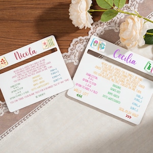 May include: Two white game cards with the names "Nicola" and "Cecilia" in cursive. Each card has colorful illustrations and text detailing game rules. The cards are on a wooden surface, with flowers and lace in the background.
