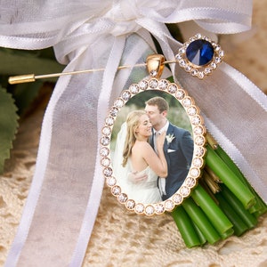 Puede incluir: Un prendedor dorado para ramo de boda con una foto de una pareja, rodeada por un marco ovalado brillante. El prendedor presenta una gema azul y está sujeto a una cinta blanca y un ramo de tallos verdes.