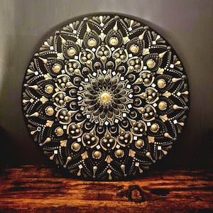 May include: A circular mandala artwork in black, adorned with detailed dot patterns in gold, grey, and white. The design showcases a central golden circle and symmetrical floral patterns. The piece is presented on a wooden surface.