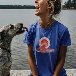 May include: A woman wearing a blue t-shirt with a red and orange sun and a wave graphic. The text "Rainy Waves" is printed on the shirt. She is laughing and looking at a dog.