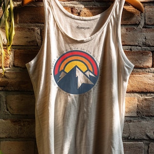 May include: A beige tank top with a round graphic featuring a mountain scene with a rainbow and the text "Spent a Little Time on the Mountain". The tank top is hanging on a wooden hanger.