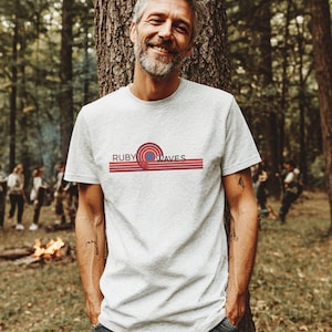 Könnte beinhalten: Ein graues T-Shirt mit einem roten, blauen und weißen Grafikdesign, auf dem "RUBY WAVES" steht.