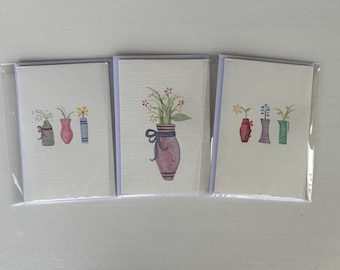 Könnte beinhalten: Drei Grußkarten in Kunststoffverpackung, jeweils mit einer Aquarellillustration einer Vase mit Blumen. Die Vasen sind in Grau-, Rosa- und Blautönen gehalten, mit verschiedenen Blumenarrangements.