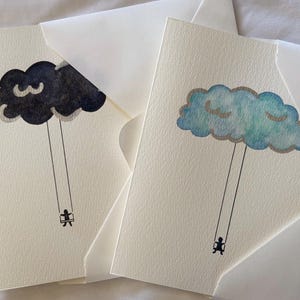 May include: Two blank greeting cards with envelopes. Each card features a watercolor illustration of a cloud with a person swinging from it. One cloud is black and the other is blue.