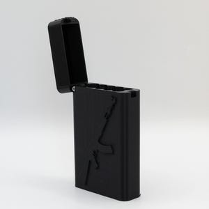 Puede incluir: Un encendedor rectangular negro con la tapa abierta, revelando el interior. El lateral del encendedor presenta un relieve detallado de un rifle. El encendedor está sobre un fondo blanco liso.