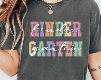 May include: A charcoal gray short-sleeve t-shirt featuring the word KINDERGARTEN printed in a blocky, multi-colored font with a white cursive script overlaying the text. The shirt has a crew neckline and a relaxed, vintage-washed appearance.