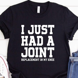 May include: Black t-shirt with white text that reads "I JUST HAD A JOINT REPLACEMENT IN MY KNEE." The shirt is paired with blue jeans and white sneakers. A small green plant is also in the image.