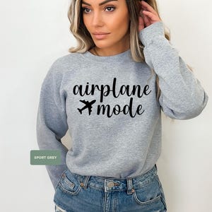 May include: Sport grey sweatshirt with the words "airplane mode" in black script and a small airplane graphic. The sweatshirt is a crew neck style and is made of a soft material. The model is wearing blue denim shorts.
