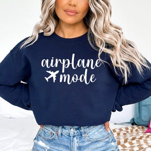 May include: Navy blue sweatshirt with the words "airplane mode" and a white airplane graphic. The text is in a cursive font. The sweatshirt is paired with blue jeans.