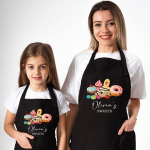 May include: Black aprons with a colorful sweets design, worn by a woman and a child. The aprons feature cupcakes, donuts, and macarons, with the text "Olivia's SWEETS". The aprons are a matching set.