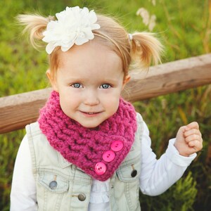 May include: A young girl with pigtails wears a pink knitted scarf with pink buttons. She is wearing a white shirt and a denim vest. She is smiling and looking at the camera.