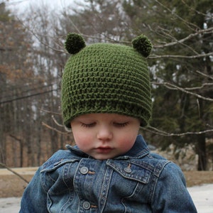 May include: A young child wearing a green crocheted hat with ear flaps, a denim jacket, and blue jeans. The child is sitting on a rock in a wooded area.