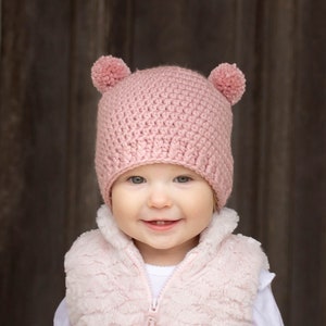 May include: A young child wearing a pink crocheted hat with ear flaps and a pink faux fur vest. The child is smiling and looking at the camera.