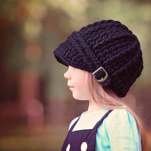May include: A black knitted beanie with a fold-down brim and a metal buckle. The beanie is worn by a young person with long brown hair.