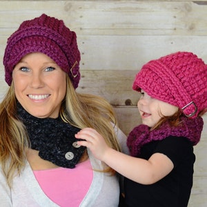 May include: A woman and a young girl wearing matching pink and purple crocheted hats with a buckle detail. The woman is also wearing a black crocheted neck warmer with buttons.